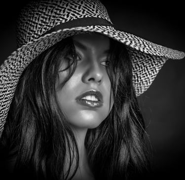 Ein Close-up-Porträt einer Frau in Schwarz-Weiß, die einen breitkrempigen Hut mit einem gemusterten Band trägt. Ihr Blick ist