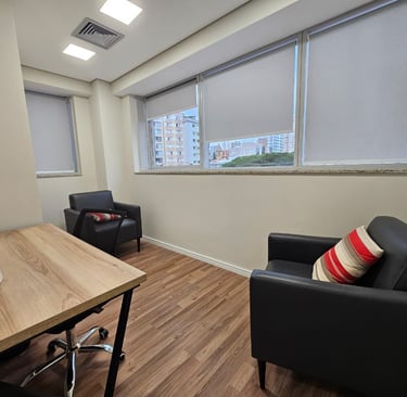 Um consultório de psicologia com duas poltronas de couro preto, uma mesa de madeira clara e uma jane