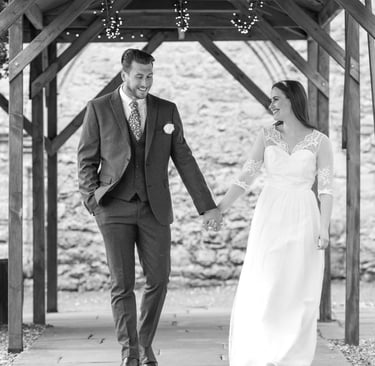 black & white photo of bride and groom walking and holding hands, both smiling, wooden canopy is framing them