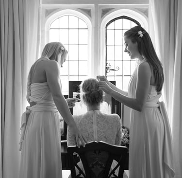 black & white photo of bridesmaids helping bride get ready, they are in front of a large window, curtains are framing them