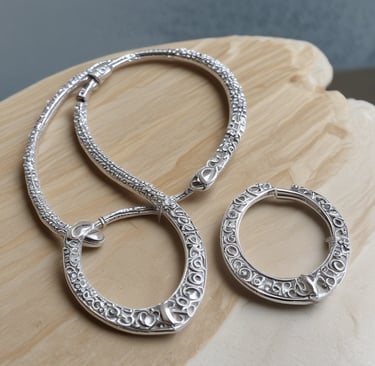 A handcrafted necklace with intricate beadwork and metal detailing lies on a textured stone surface. Surrounding the necklace are delicate yellow and pink flowers with long green stems and leaves. The overall appearance is both rustic and elegant, blending natural elements with artisan jewelry.