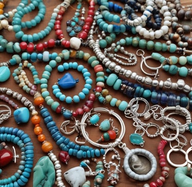 An assortment of jewelry is arranged on a dark surface with vibrant green leaves. The collection includes necklaces with intricate designs featuring gemstones. Notable pieces include a spider-shaped pendant with a white gemstone and a necklace with a large blue stone surrounded by smaller pearls. Another item is a silver pendant with branch-like elements. The background contrasts with dried leaves adding a natural element.
