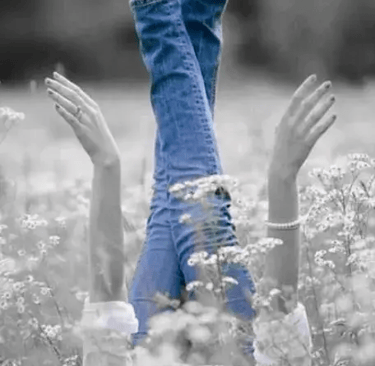 A black and white image of a woman kicking her legs up in a field. The only color is her blue jeans.