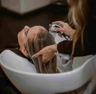 A hair stylist washes a blonde woman's hair in a white salon sink basin using a professional sprayer.