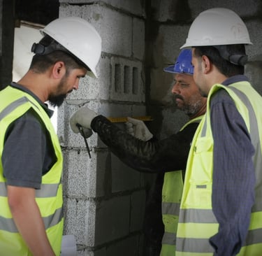 Construction workers in hard hats and safety vests measuring a cinder block wall at a building site.