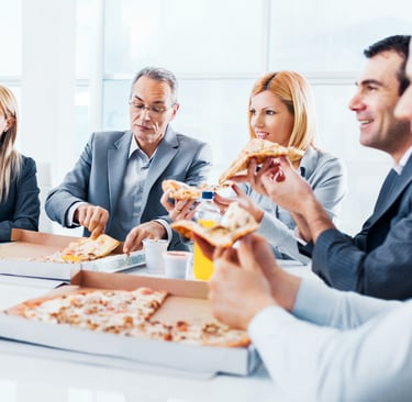 مجموعة من الموظفين يرتدون بدلات رسمية يتناولون البيتزا معًا خلال استراحة غداء عمل في مكتب حديث مضاء
