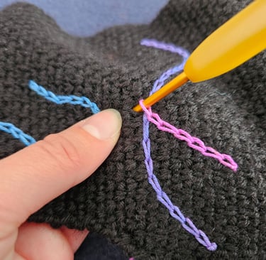 A yellow crochet hook making surface slip stitches with colorful yarn on black fabric.