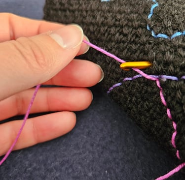Hand holding pink thread and golden crochet hook to embroider details on a black crochet fabric.