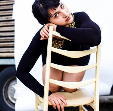 Eine junge Frau in einem schwarzen Kleid hockt auf einem Stuhl.