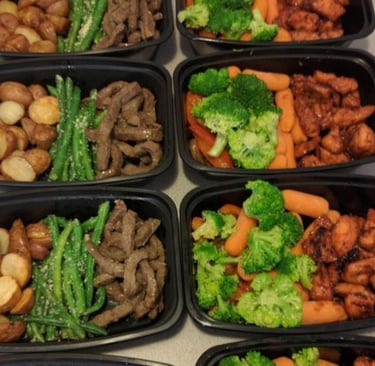 Meal prepped food in black Tupperware including steak, carrots, broccoli, asparagus and potatoes