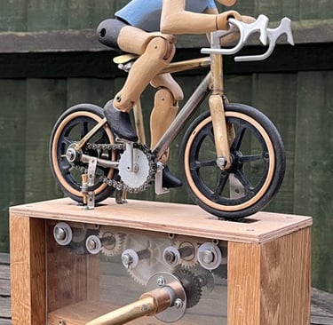Articulated carved wooden cyclist on brazed metal miniature road bike, on base with visible cogs