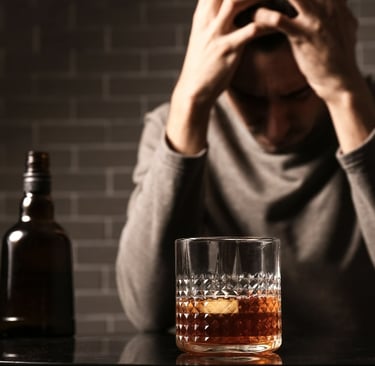 A distressed man with his head in his hands sits at a table in front of a bottle and a glass of liquor