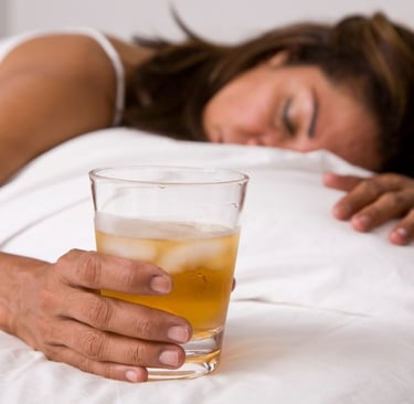 A woman looking unwell lies in bed while holding a glass containing an alcoholic drink, depicting the negative consequences