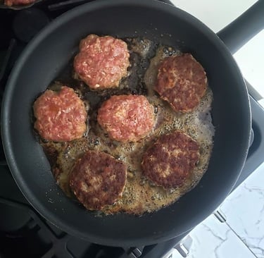 Sizzling ground beef burger patties frying in a non-stick skillet on a gas stovetop.