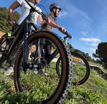 Two mountain bikers in helmets standing on a grassy trail with their mountain bikes on a sunny day.