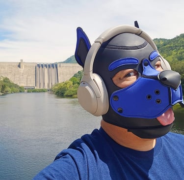 Man in a pup mask wearing headphones standing on a river bank near a large dam.