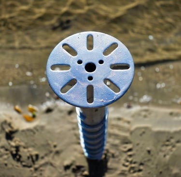 A light blue circular metal screw pile foundation cap installed over beach sand.
