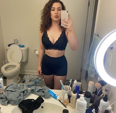 Woman in a black lace bralette and high-waisted shorts posing for a mirror selfie in a bathroom.