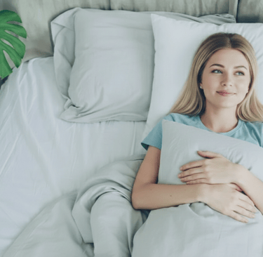 A smiling young woman lying in a comfortable bed with white pillows and a monstera plant.
