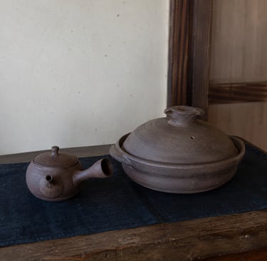Handmade brown ceramic Japanese kyusu teapot and donabe clay pot on a rustic wooden table.