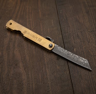 A Japanese Higonokami folding knife with a Damascus steel blade and brass handle on a dark wood surface.