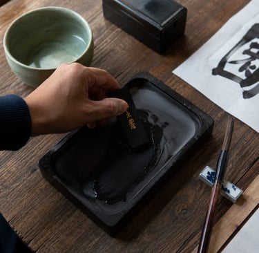 A hand grinds a traditional black ink stick on an inkstone for Chinese calligraphy.
