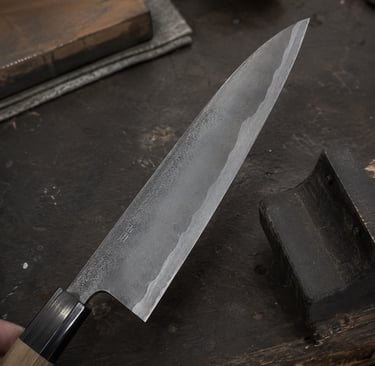 Hand-forged Japanese Gyuto chef knife with a carbon steel blade on a rustic wooden sharpening bench.