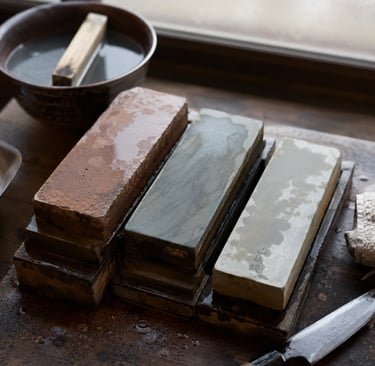 Professional Japanese whetstones in various grits for sharpening a kitchen knife on a wooden workbench.