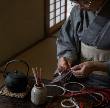 A Japanese woman in a grey kimono practicing Mizuhiki paper cord art next to a cast iron teapot.