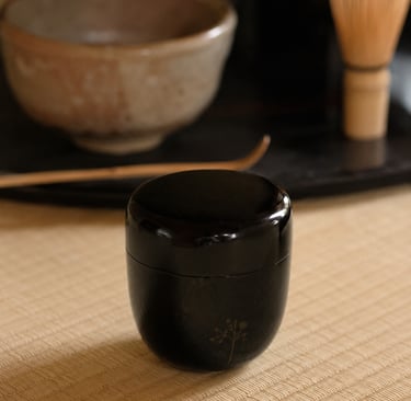 A black lacquer natsume tea caddy on a tatami mat with a matcha bowl and bamboo whisk.