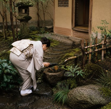 A woman in a traditional Japanese kimono performs a purification ritual at a stone water basin in a mossy garden.