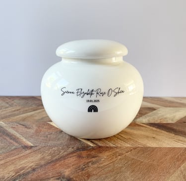 Rounded white ceramic urn with glossy finish, black script text “Sienna Elizabeth Rose O'Shea” and a small rainbow con.