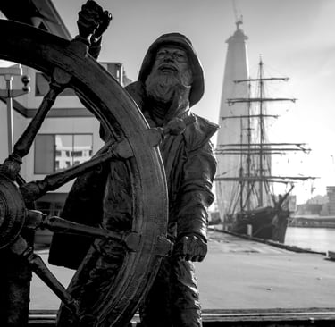 Black and white statue of a sailor at a ship's wheel overlooking a harbor with a tall ship.
