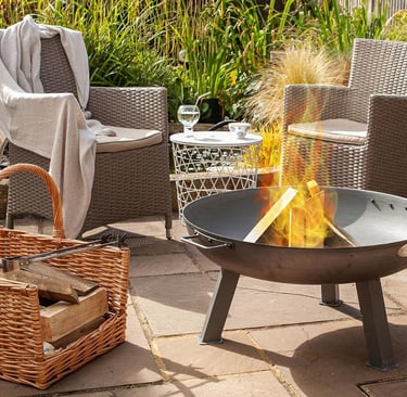 A portable steel fire pit burning logs on a stone patio with wicker garden chairs and a wood basket.