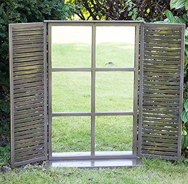 Rustic garden mirror with shutters reflecting a green lawn to create a window effect in a backyard.