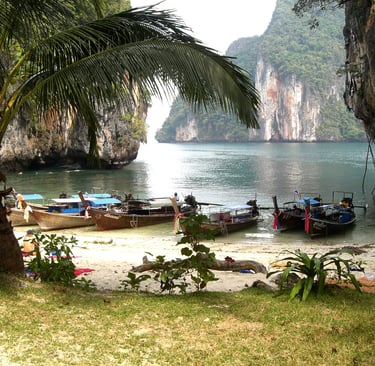 Traditional Thai longtail boats anchored at a tropical beach with limestone cliffs and palm trees