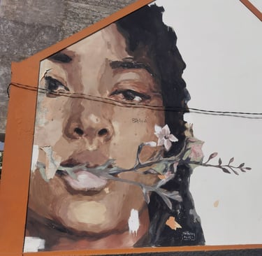 Mural painting of a woman's face with a flower branch in her mouth on a white building wall.