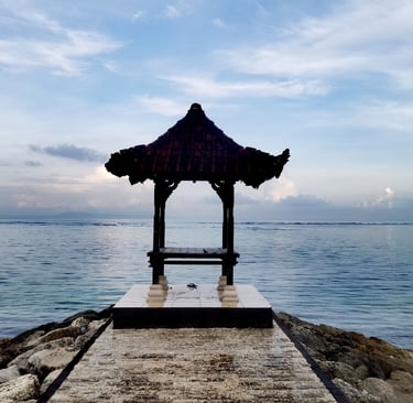 Traditional Balinese ocean gazebo on a stone pier overlooking the blue sea under a cloudy sky.