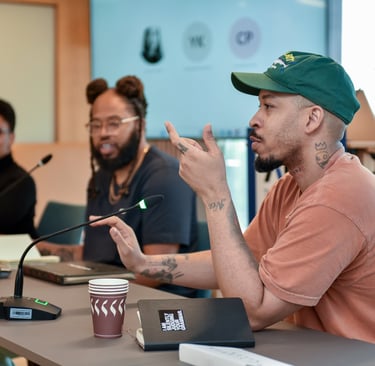 Un conférencier américain est en train de parler face à un micro, assis à côté de ses collègues.