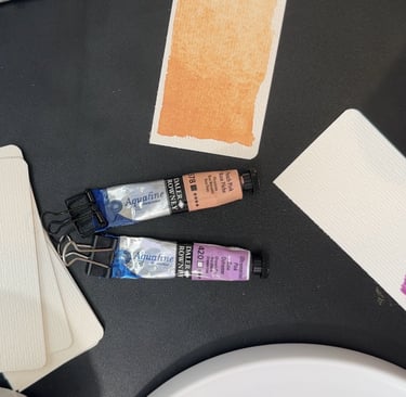 Watercolor paint tubes, watercolor paper, and someone painting on a piece of paper. black background