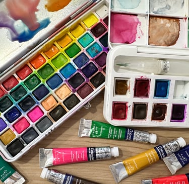 Overhead view of watercolor paint palettes, paint tubes, and brushes arranged on a wooden table — essential supplies for lear
