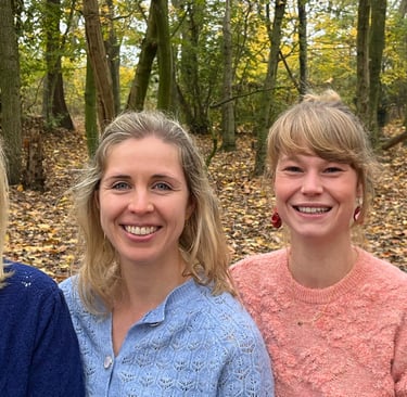 Four smiling women standing together in a scenic autumn forest with colorful fallen leaves.