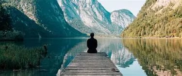Homme de dos assis sur un ponton face à un lac ecaissé entre deux montagnes
