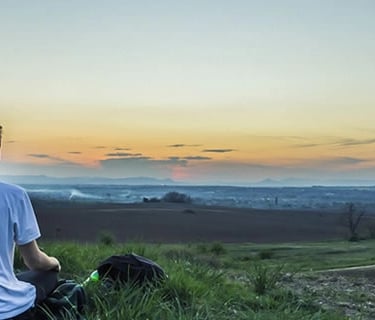 Jeune homme de dos assis dans l'herbe admirant un paysage lointain et un coucher de soleil