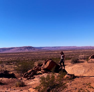 A hiker stands on a desert rock overlook with Arizona canyon mountains under a clear blue sky.