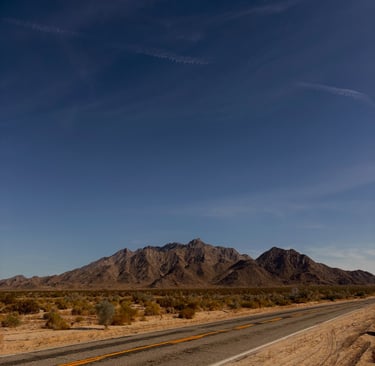 A California desert highway leading toward rugged mountains under a clear blue sky.