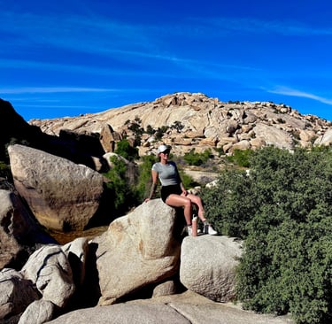 A woman sits on desert boulders in Joshua Tree National Park under a clear blue sky.