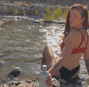 A woman in a red bikini sits by a sunlit river holding a bottle of hyaluronic acid skincare.