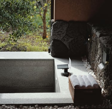 Luxury indoor-outdoor stone soaking tub with rolled white towels overlooking a lush green garden.