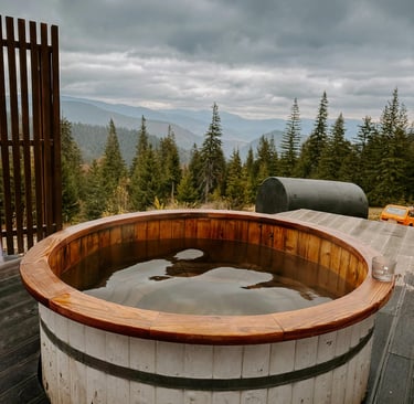 Rustic wood-fired hot tub on a deck with a scenic mountain and forest view under a cloudy sky.